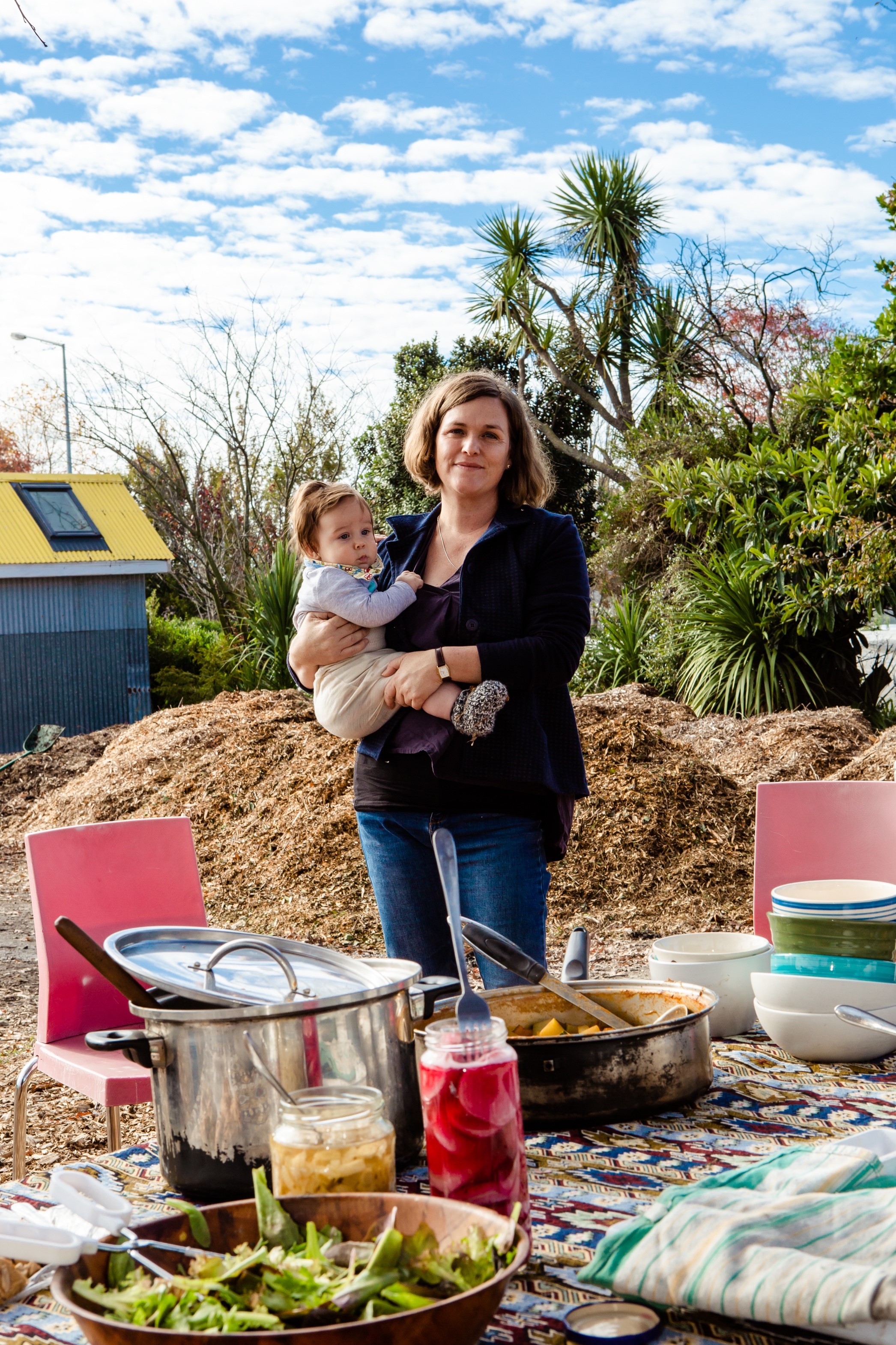 Kelly stands holding a baby. Behind her is a pile of mulch and an urban garden. In front, a table laid out with food. 