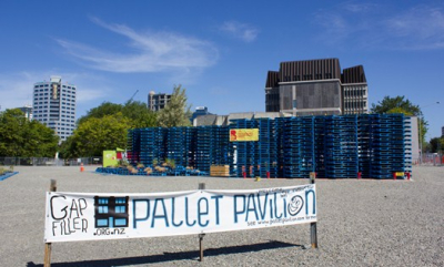 A structure made of pallets is in the background. In front a sign reads "Gap Filler Pallet Pavilion". In the distance two tall buildings can be seen, while in the foreground it is mostly gravel. 
